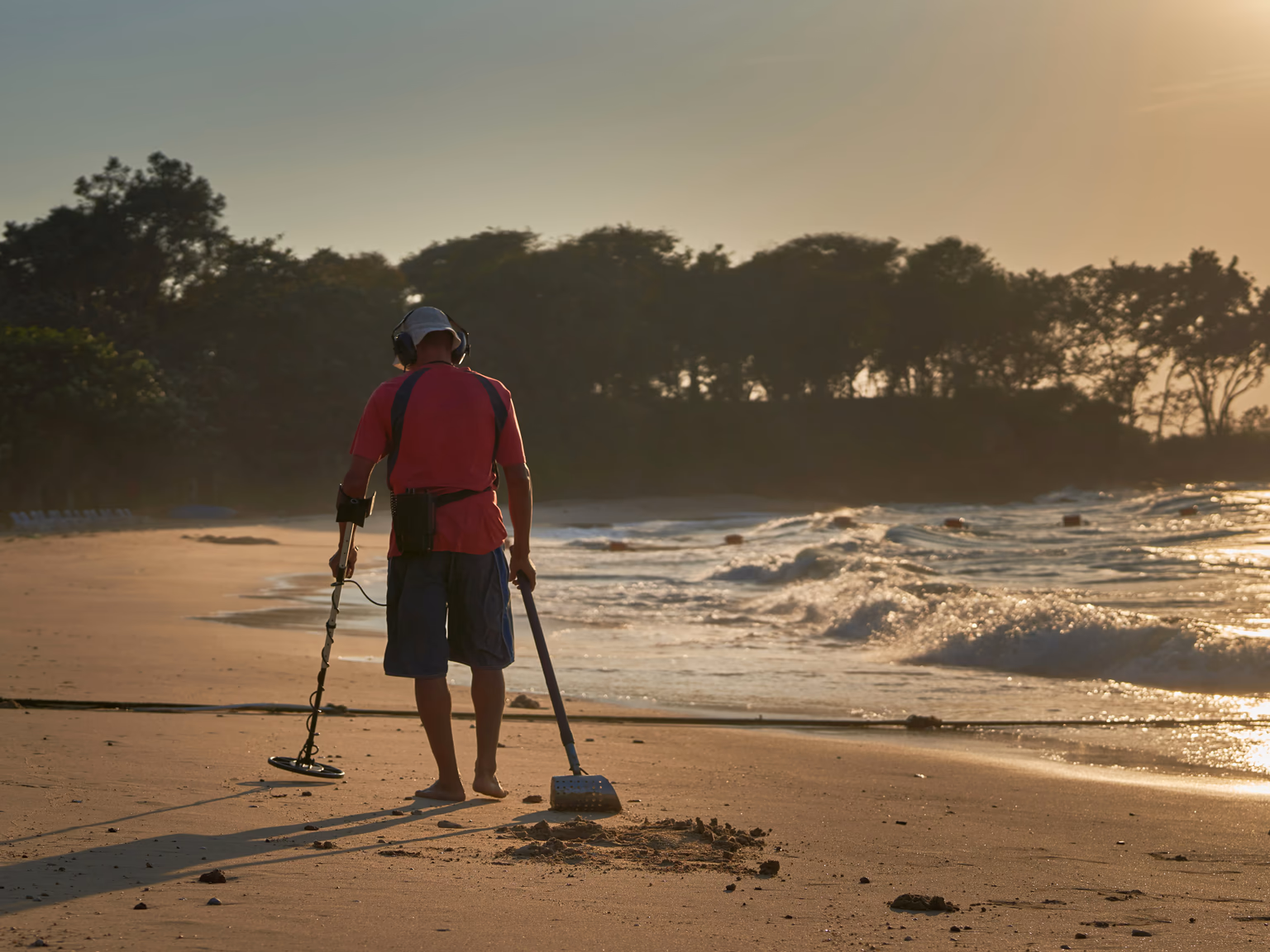 Sondengänger mit Kopfhörern und Sandschaufel sucht bei Sonnenuntergang den Spülsaum eines Strandes ab