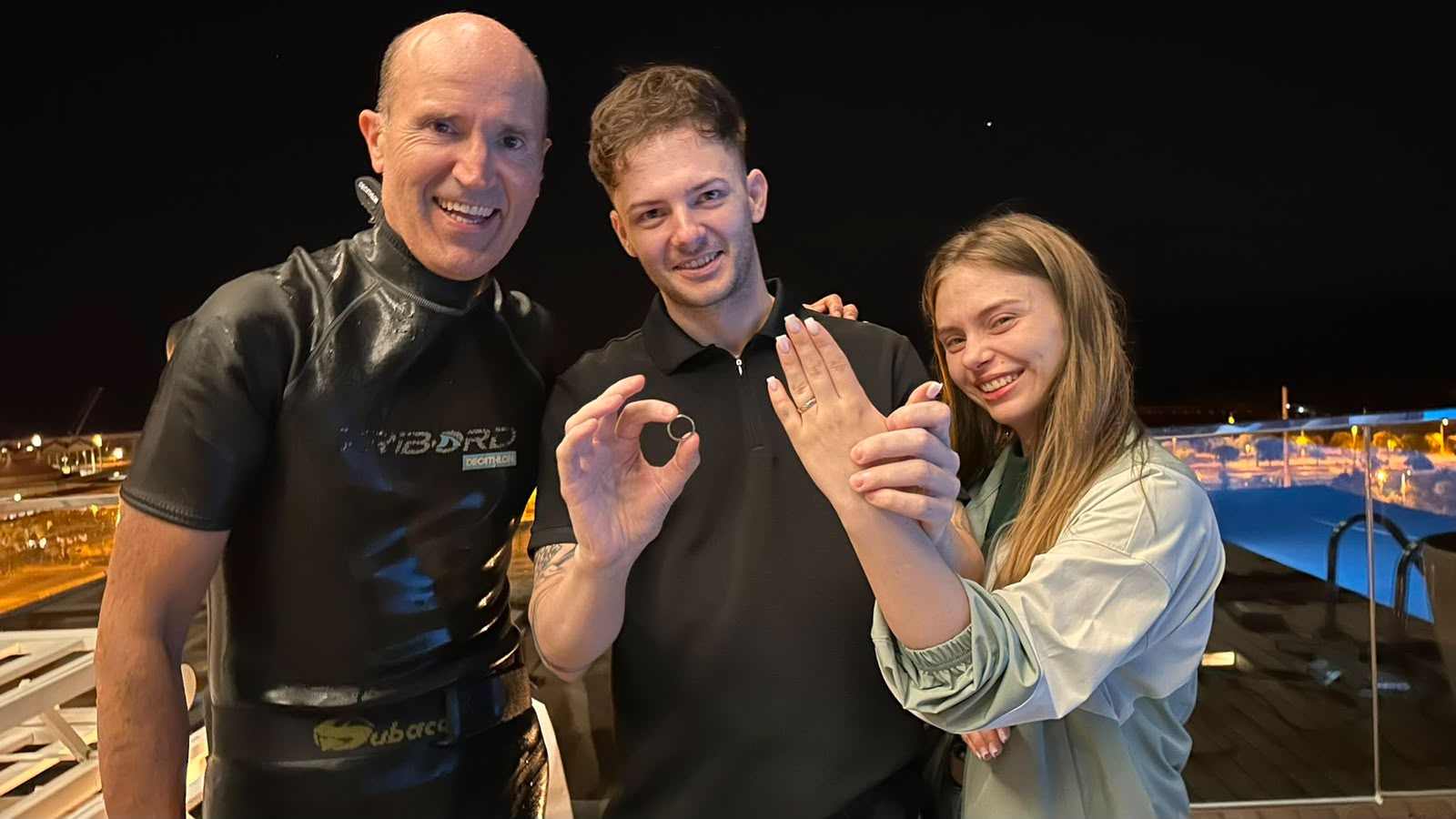 Radu holding up the recovered ring and his wife showing her hand, next to our detectorist in a wetsuit, on the hotel rooftop in Barcelona at night with the pool behind them