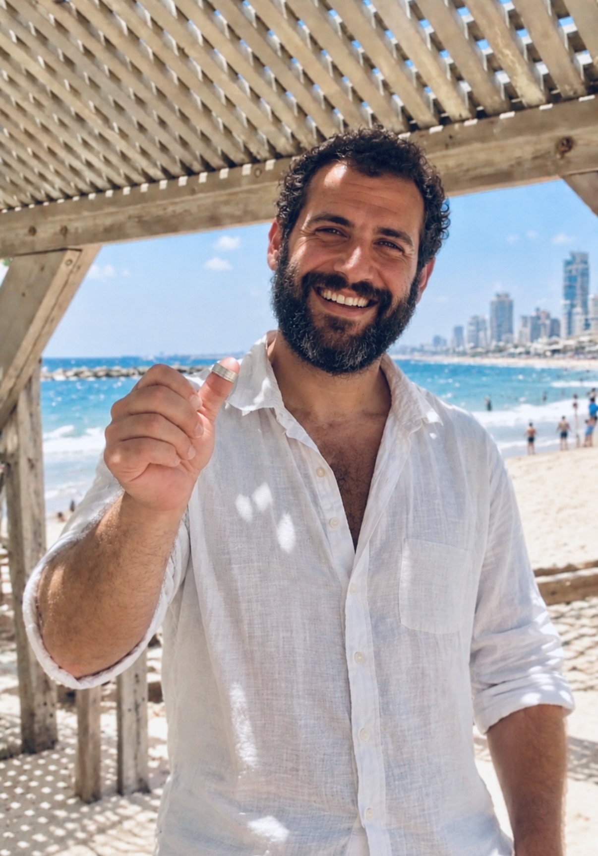 The smiling owner on Jerusalem Beach in Tel Aviv, holding up the recovered white gold wedding ring on his thumb, with the sea and the city skyline behind him