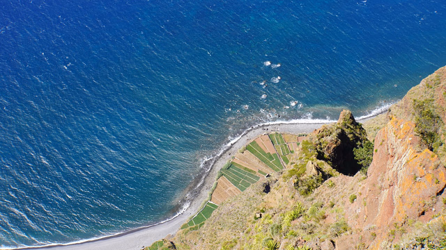 Dark volcanic pebble beach below green cliffs on Madeira with blue Atlantic water, the kind of shore where a lost ring slips down between the stones